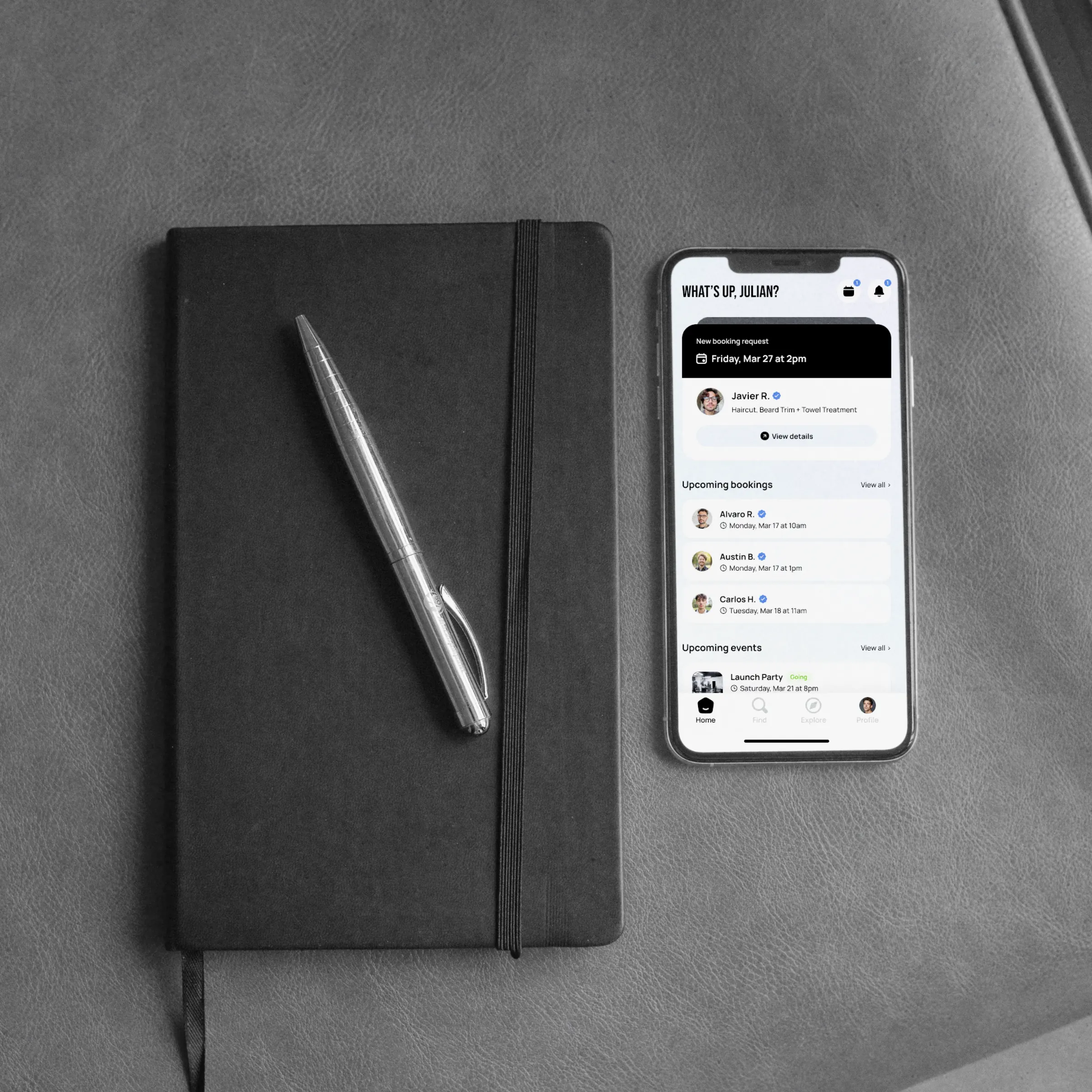 A phone on a leather desk beside a notebook and pen, showing Empire Biz — a new booking request from a client, followed by the barber&rsquo;s upcoming bookings and events